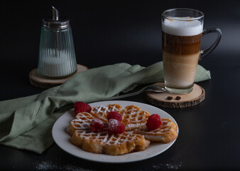 A freshly baked waffle with raspberries and powdered sugar on a white plate and Kaffe Latte Macciato