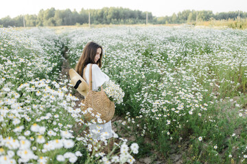 portrait of a beautiful young Caucasian woman on a chamomile field in a picturesque valley. a young lady in a white cotton dress and a straw hat. holds a bouquet of daisies. 
