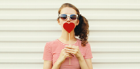 Portrait close up of beautiful young woman blowing her lips with red lipstick with red sweet heart shaped lollipop on white background