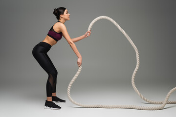 side view of sportswoman training with battle ropes on grey background.