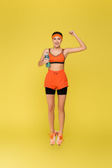 cheerful sportswoman with sports bottle jumping and showing win gesture isolated on yellow.