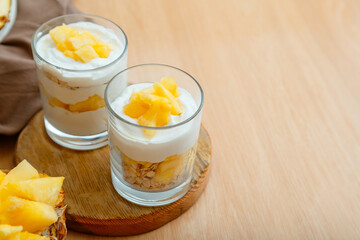 Obraz na płótnie Canvas Tasty pineapple desserts with chopped fresh juicy pineapple. Breakfast dessert with oat granola, greek yogurt and pineapple in layers in glass on wooden table with copy space