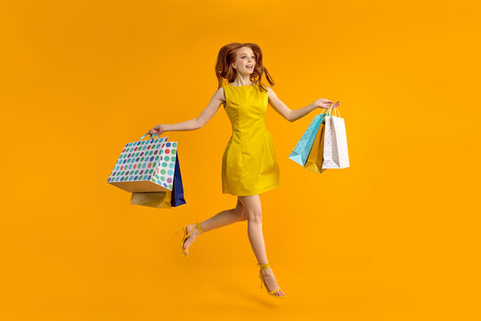 Full length image of charming young redhead woman wearing yellow dress smiling, caucasian lady is carrying colorful paper shopping bags, jumping and looking away isolated over yellow studio background