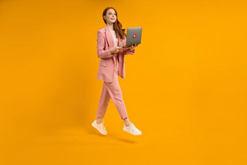 Cheerful jumping female with laptop isolated on yellow studio background. Modern technologies,...