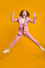 Fototapeta na wymiar Excited redhead successful business woman jumping with raised fingers showing up isolated on yellow background, wearing pink suit, shocked surprised, emotionally reacts on something