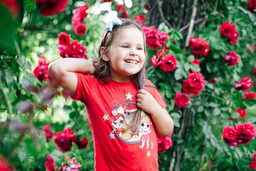 close-up portrait of a positive, laughing girl in a red T-shirt playing with her hair in a garden with red bush roses.