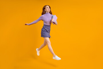 Fototapeta na wymiar beautiful redhead lady at weekend jumping, hold notebooks in hands on way home, in air, wearing casual shirt and skirt isolated yellow orange background. Side view, copy space. Full-length portrait