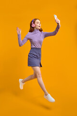 Fototapeta na wymiar Excited Redhead Woman In Heaphones Jumping, Walking And Talking On Phone Online Via Camera, Say Hello, Smiling, Isolated On Yellow