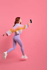 side view portrait of redhead female in leggins running jumping forward with smartphone in hands, taking photo of something, hurry. isolated pink background