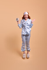 Emotional redhead woman isolated on beige background in pajamas, holding clock with surprised facial expression, jumping. Eyemask on forehead, female look at camera, portrait. copy space