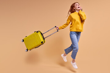 Traveler tourist redhead woman with suitcase, jumping in casual outfit isolated in studio. Caucasian Female passenger traveling abroad on weekends. Air flight journey concept. Copy space