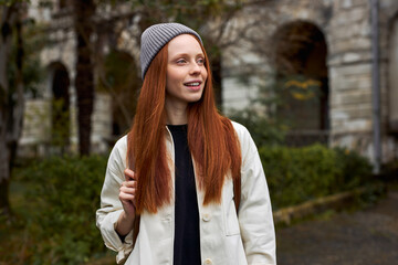 Close-up Portrait Of Dreamy Woman With Natural Red Hair On Travel Trip, Outdoors, Posing At Camera, Smiling, in Coat And Hat. Old Building In The Background © alfa27