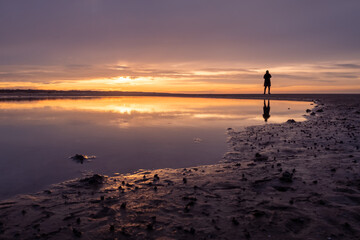 a sunset over a beach with a person standing at the horizon. reflections in the water
