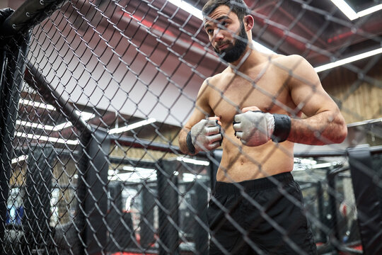 Portrait of MMA fighter thinking about strategy for match, standing on ring preparing for fight, shirtless muscular male is going to practice fighting. shot through cage