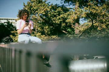 An attractive young woman with curly hair sitting outdoors with headphones on and choosing music on the phone.