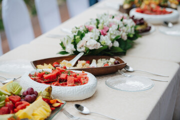 Variety of fuits and meats served on a big narrow dining table. Wooden, glass and ceramic dishes layed in a straight line.