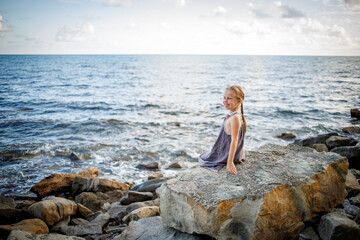 A blonde girl in a sundress sits on a stone on the seashore.