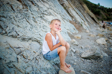 A boy in a white T-shirt and denim shorts sits on a rock near the seashore