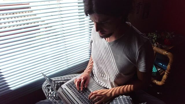 Young freelancer man in bed homeoffice in gray striped pajamas studying working with a laptop on his legs. Sunlight in the blinds 