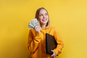 portrait of young exciting smiling pretty woman holding a stack of money dollar bills and laptop. Teenager Girl dreaming of money, earnings online,lottery win isolated on yellow color background