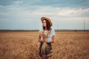 Girl in a straw hat stands in a wheat field with a bouquet