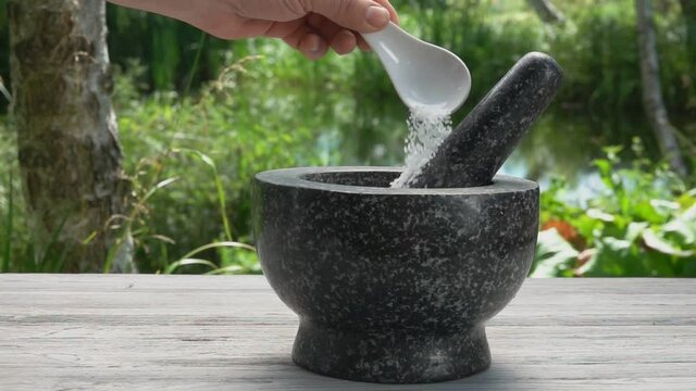 Hand is putting white sea salt in the grey stone mortar in slow motion outdoors