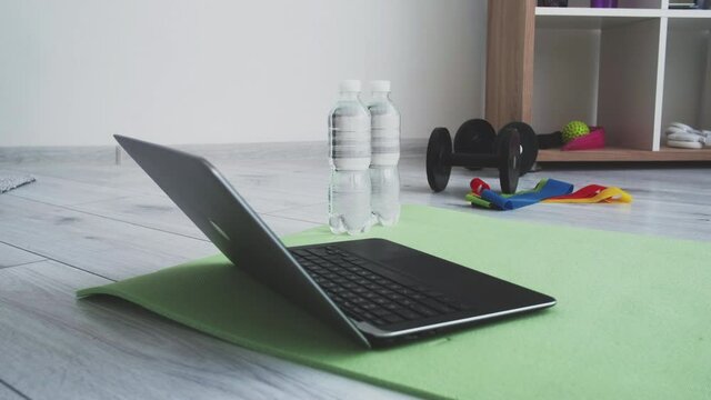 Sport equipment. Home fitness. Online training. Healthy lifestyle. Light room with laptop on green yoga mat dumbbells resistance bands bottles of water on floor motion view.