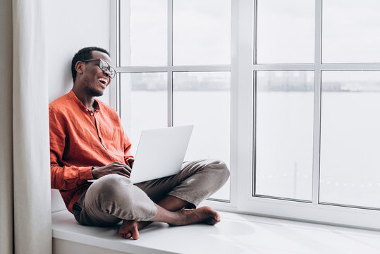 Joyful African-American guy in stylish clothes laughs at video call via modern laptop sitting on large window sill at home