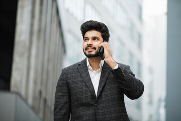 Smiling hindu man in business suit standing in street with suitcase in hands and talking on mobile phone. Concept of people, work and communication.