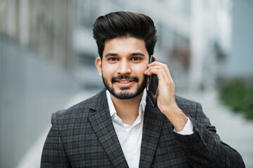 Portrait of positive arabian man in formal wear having mobile conversation while standing on city street. Blur background of modern office building. Concept of business and technology.