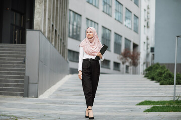 Full length portrait of arabian woman in hijab standing near modern business center with smartphone and clipboard in hands. Successful business lady posing outdoors.