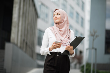 Low angle view of beautiful muslim woman standing near modern office building with smartphone in...