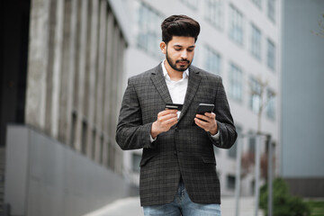 Handsome hindu man in stylish formal clothes using modern smartphone and credit card for online shopping. Bearded businessman standing on street and making purchase.
