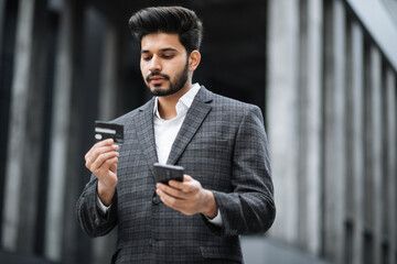 Stylish bearded man doing online purchases while standing near office building. Muslim guy in formal wear using credit card and smartphone for shopping.