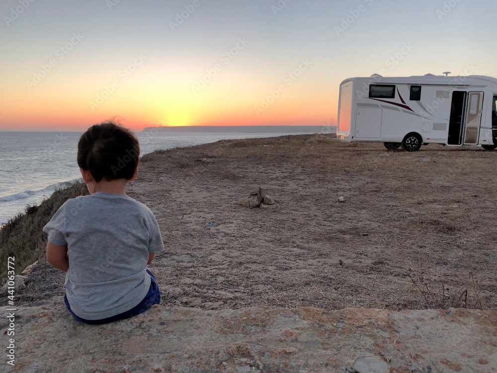 Wall mural Child on his back sitting looking at the sunset by the sea near a caravan.