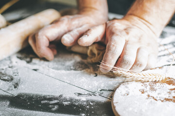 Male hands knead dough with flour next to clay pot and oil bottle and rolling pin on dark table, while cooking