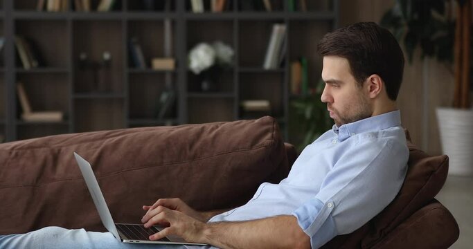 Serious focused millennial man seated in comfy sofa with laptop, typing on netbook keyboard, make business remotely from home side view. Telecommuting activity, modern digital technology usage concept