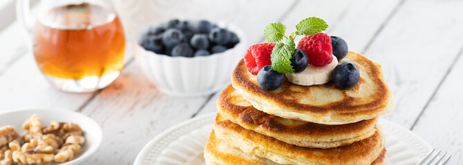 Narrow view of a stack of buttermilk pancakes topped with berries and banana slices and served with syrup and walnuts.