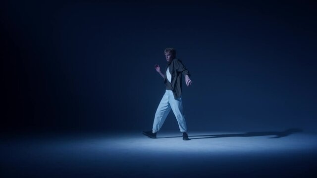 Young blonde guy dancing modern choreography in dark studio background. Talented choreographer man showing the new hip hop movements. Shooting a dance video clip. Hip hop school for teenagers. 
