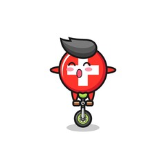 The cute switzerland flag badge character is riding a circus bike