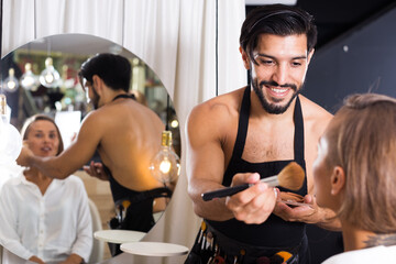 Cheerful male makeup artist in salon applying cosmetics for woman