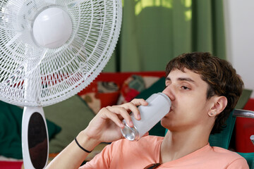 Concept of summer at home. Young man on a beach chair, drinking beer or soft drink, near the fan.