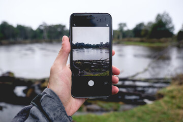 Hand holding a phone with the view (picture in picture) of a lake, trunks and forest in the Chilean patagonia in a rainy day