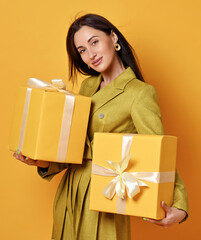 Portrait of young smiling slim brunette woman in yellow skirt and short jacket holding in hands two yellow gift boxes, birthday presents with bows, carrying over yellow background