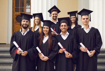 Group of happy students in mortarboards and bachelor gowns with diplomas celebrating success on university campus. Education, graduation and people concept.