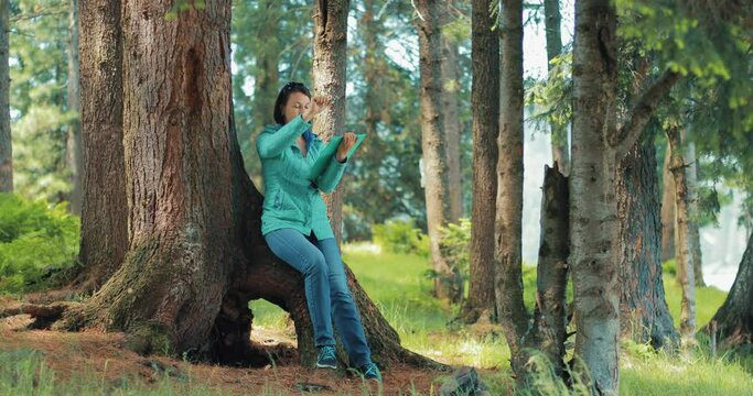 A girl draws in the forest. She is sitting on the root of a large tree and holds a paper stand in her hand. She has a pencil in her right hand. A girl draws a landscape.