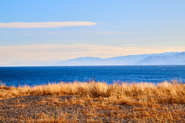 Beautiful natural view of the landscape with the shore with yellow grass, lake, blue sky and mountains or hills on background with white clouds on a sunny autumn day