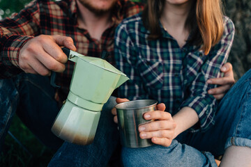 Close-up of a tourist guy pours a girl coffee or tea into a mug from a geyser coffee maker. Travel to the forest to drink drinks. Hot coffee from a cup. Family on vacation after walking.