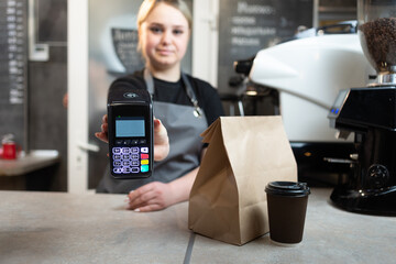 Contactless payment of an order through a payment terminal with nfs to a barista girl in a cafe for coffee and food in paper packaging.The girl holds out the device to pay for the order.