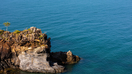 Orange brown rock cliff with calm sea waves during a sunny day at a tropical rocky beach with green plants foreground. Rocky island, a destination for tourists in summer time